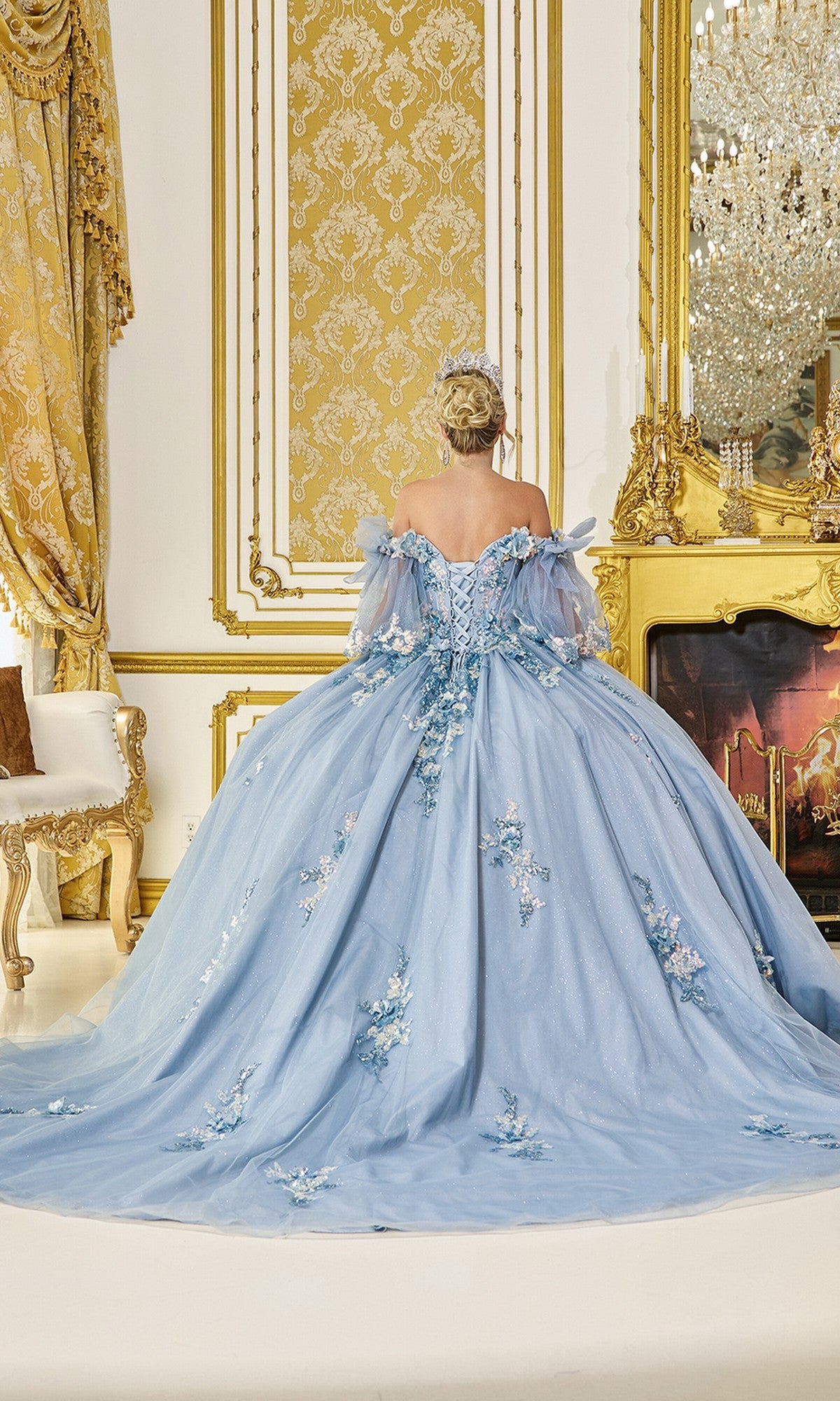 A woman wearing the Quinceanera Dress 5072 By Dancing Queenโa blue ball gown with floral accents and glitter tulleโstands facing a gold-adorned wall and fireplace in a luxurious room.